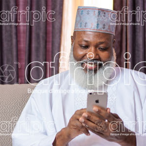 Homme africain senior souriant, assis dans son salon, et manipulant son téléphone