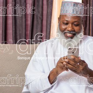 Homme africain senior souriant, assis dans son salon, et manipulant son téléphone