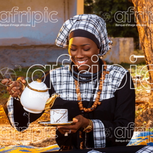 Jeune femme africaine en tenue traditionnelle toute souriante en période de Magal, assise à l'extérieur sur une natte et servant du café