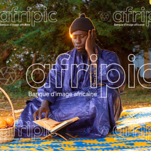 Jeune africain en tenue traditionnelle, assis sur une natte à l'extérieur et lisant le coran.