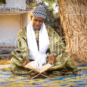 Jeune africain en tenue traditionnelle, assis sur une natte à l'extérieur et lisant le coran.