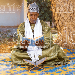 Jeune africain en tenue traditionnelle, assis sur une natte à l'extérieur et lisant le coran.
